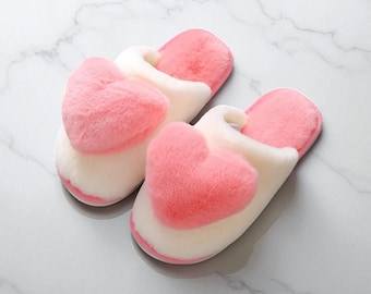 Può includere: Un paio di pantofole soffici con un design a forma di cuore rosa. Le pantofole sono bianche con dettagli a forma di cuore rosa. Il materiale morbido e felpato suggerisce comfort. Il design è semplice e carino.