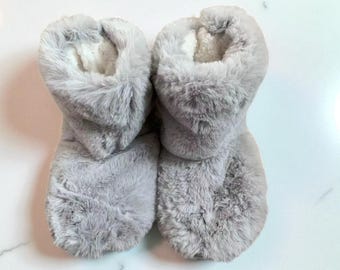 May include: A pair of soft, gray faux fur slipper boots. The boots have a fluffy exterior and a white, plush interior. The boots are designed to cover the feet and ankles, offering warmth and comfort. The slippers are ideal for indoor use.