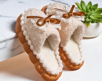 Può includere: Un paio di pantofole color crema con interno soffice e suola marrone strutturata. Ogni pantofola presenta un bordo in pizzo bianco decorativo e un fiocco in raso marrone. Le pantofole sono progettate per l'uso in casa.