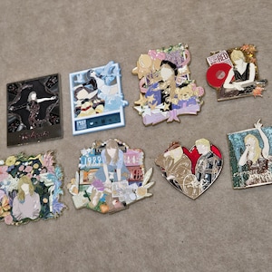 Puede incluir: Una colección de ocho pines de esmalte decorativos que presentan diversas ilustraciones artísticas de figuras, patrones florales y temas musicales. Los pines varían en forma, incluyendo diseños rectangulares, en forma de corazón e irregulares con colores vibrantes y acentos metálicos.