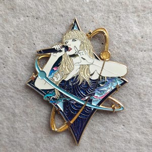 Op de afbeelding: Een goudkleurige emaille speld met een blonde figuur die in een microfoon zingt. De figuur draagt een donkerblauwe glitterjurk tegen een ruitvormige achtergrond met gouden orbitale ringen, blauwe accenten en kleine stervormige details.