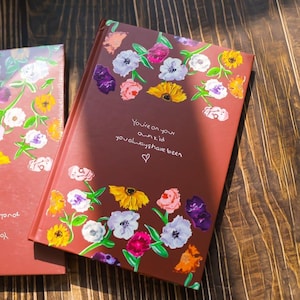 May include: A maroon journal with a floral design featuring white, yellow, pink, and purple flowers. The cover has the text "You're on your own kid, you always have been" with a heart symbol.