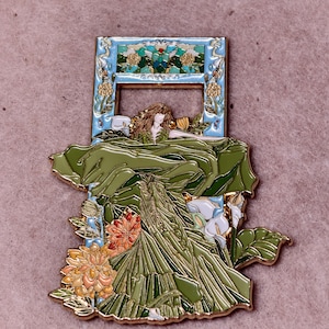 Puede incluir: Un broche de esmalte y metal dorado que muestra una figura de cabello largo castaño con un vestido verde fluido. El diseño incluye hojas grandes verdes, calas blancas y flores naranjas, enmarcado por un borde rectangular azul claro con detalles florales.