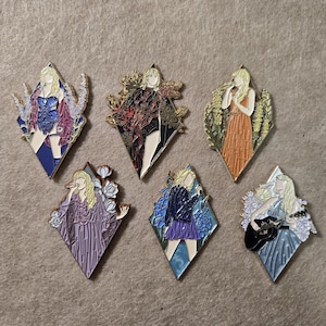 Puede incluir: Un conjunto de seis pines de esmalte en forma de diamante que muestran figuras estilizadas con cabello rubio. Cada pin representa un atuendo diferente y un fondo floral o botánico, con esquemas de color en azul, negro, naranja y violeta.