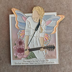 Peut inclure: Une broche rectangulaire en émail représentant une silhouette aux cheveux blonds tressés, vêtue d'une robe blanche et tenant une guitare. La silhouette a de grandes ailes scintillantes multicolores et est entourée de roses roses. Le texte indique : Our song is the way he laughs, The first date man I didn't kiss him and I should have.