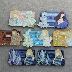 Peut inclure: Une collection de sept broches en émail rectangulaires en forme de cassettes audio, chacune avec un coloris unique, des éléments floraux et une silhouette stylisée. Les motifs incluent des textes comme Red, 1989, Fearless et Midnights.