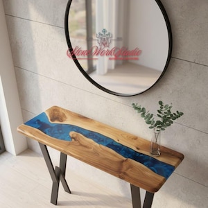 Puede incluir: Una consola de madera con un diseño de río de resina azul. La mesa tiene patas de metal oscuro y un jarrón de vidrio transparente con vegetación. Un espejo redondo con marco negro cuelga de la pared encima de la mesa.