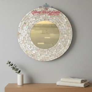 May include: Circular mirror with a mosaic-style frame, made of small, rectangular, off-white tiles. The mirror has a gold-toned centre and is mounted on a grey wall. A wooden shelf below holds a vase and folded towels.