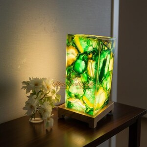 May include: A rectangular lamp with a vibrant green and yellow stone design. The lamp sits on a wooden base and is illuminated, showcasing the colors. A small bouquet of white flowers is to the left.