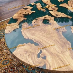 Pu&ograve; includere: Tavolo rotondo in legno dal design unico. Il tavolo presenta un motivo a venature di legno marrone chiaro con sezioni di resina verde scuro. I bordi del tavolo sono marrone scuro. Le parole "STONE WORKSTUDIO" sono stampate.