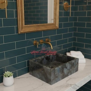 Op de afbeelding: Een rechthoekige, donkergrijze stenen wastafel met blauwe en grijze adering staat op een wit aanrechtblad. De wastafel staat in een badkamer met tealkleurige metro tegelwanden, een goudomrande spiegel en gouden armaturen. Een kleine vetplant in een witte pot staat op het aanrecht.