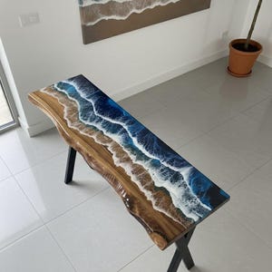 Puede incluir: Banco de madera con diseño de borde vivo, con una parte superior de resina que imita las olas del océano. El banco tiene una base de madera marrón y patas de metal negro. La parte superior de resina incorpora tonos de azul, blanco y marrón.