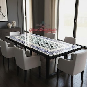 May include: A rectangular dining table with a mosaic tile top featuring a geometric pattern in shades of blue, green, and white. The table has a black frame and is surrounded by six white upholstered chairs. The room has large windows and dark wood flooring.