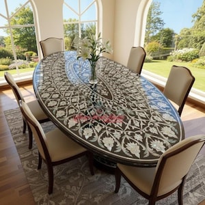 Op de afbeelding: Een ovale eettafel met een donkerbruin oppervlak ingelegd met een bloemmotief in wit en lichte kleuren. Zes beige stoelen omringen de tafel. Een vaas met witte bloemen staat in het midden. De tafel staat in een kamer met grote ramen.