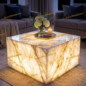 Può includere: Tavolino da caffè quadrato illuminato in marmo con venature dorate. Un vaso di fiori e una candela accesa sono sul piano. L'ambiente, con arredi dai toni neutri, crea un'atmosfera calda ed elegante. Dimensioni: circa 100cm x 100cm.