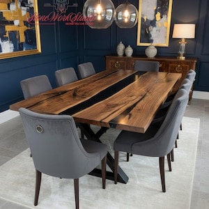 Puede incluir: Una gran mesa de comedor de madera con una incrustación de resina epoxi oscura, rodeada de ocho sillas grises tapizadas. La mesa está sobre una alfombra clara en una habitación con paredes azul oscuro y obras de arte con marcos dorados. Dos lámparas colgantes de globo de cristal cuelgan encima.