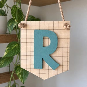May include: A light-coloured wooden pennant sign featuring a black grid pattern and a large, raised, teal-coloured letter R in the centre. The sign is suspended by a thick, natural-toned twisted rope knotted through two holes at the top corners.