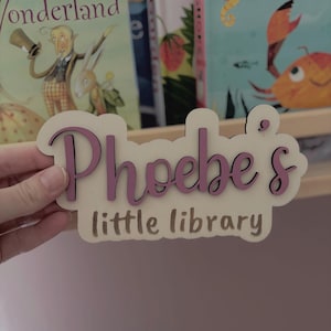 May include: A light-coloured wooden sign with a wavy, cloud-like border. The name Phoebe's is written in raised, dark purple script lettering, while the words little library are engraved in a simple, dark brown sans-serif font below the name.