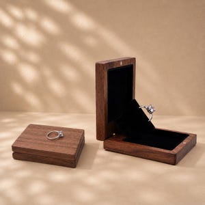 Pode incluir: Uma caixa de anel retangular de madeira castanho-escura com tampa articulada aberta, revelando um forro interior de veludo preto. Um anel de prata com uma pedra transparente é exibido dentro da caixa, enquanto um segundo anel idêntico repousa sobre a tampa.