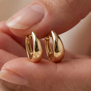 May include: A pair of small, polished gold hoop earrings with a smooth, rounded teardrop shape. The earrings feature a reflective metallic surface and a hinged closure mechanism, showcasing a simple, curved design that catches the light.