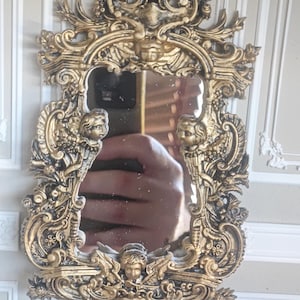 Puede incluir: Un espejo de pared de color dorado con un marco ornamentado que presenta volutas intrincadas, motivos florales y tres figuras de querubines esculpidas situadas en la parte superior, los lados y la base. Posee una superficie de cristal central.