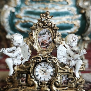 Può includere: Un orologio decorativo con cornice dorata e volute ornate, due figure di cherubini bianchi che suonano strumenti ai lati e un pannello ovale centrale con una scena dipinta sopra un quadrante rotondo con numeri romani neri.
