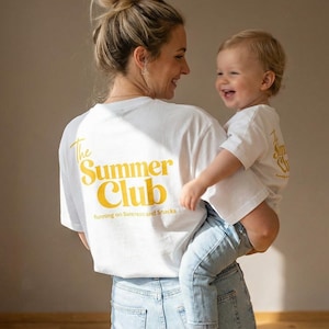 Könnte beinhalten: Eine Frau hält ein Kind, beide tragen weiße T-Shirts mit dem gelben Text "The Summer Club". Die Frau trägt eine hellblaue Jeans. Das Kind trägt eine hellblaue Jeans. Der Hintergrund ist neutral.
