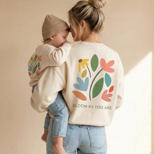 Pode incluir: Uma mulher segura uma criança, ambas vestindo moletons creme combinando com um design floral. Os moletons têm um gráfico de flores coloridas nas costas, com as palavras "BLOOM AS YOU ARE" impressas abaixo. A criança usa um gorro bege e jeans azul.