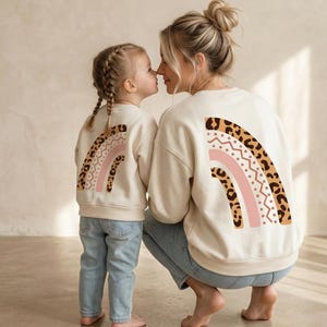 Può includere: Felpe color crema abbinate con un design arcobaleno sul retro. L'arcobaleno presenta motivi rosa, marroni e maculati. Una donna e una bambina indossano le felpe e jeans blu.