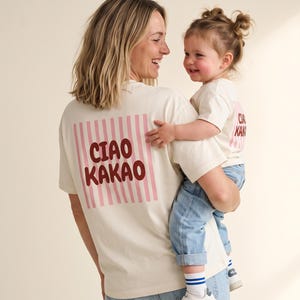 Könnte beinhalten: Zwei cremefarbene Kurzarm-T-Shirts mit einem rosafarbenen, vertikal gestreiften Quadrat auf der Rückseite und dem Schriftzug CIAO KAKAO in Dunkelrot. Die T-Shirts werden mit hellblauen Jeans und weißen Turnschuhen mit blauen Akzenten kombiniert.