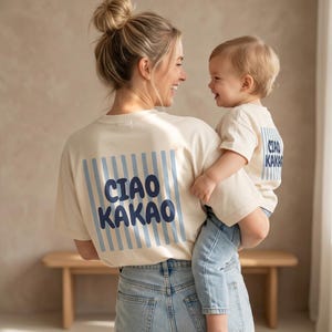 Peut inclure: Deux t-shirts de couleur crème avec un motif carré à rayures verticales bleu clair au dos. Le texte "CIAO KAKAO" est imprimé en lettres grasses, arrondies et bleu foncé sur les rayures des deux t-shirts. Les t-shirts ont des manches courtes.