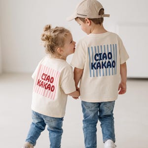 Könnte beinhalten: Zwei cremefarbene T-Shirts mit blauen Jeans. Ein Shirt hat ein rosafarbenes Streifenquadrat mit dem Text CIAO KAKAO in Rot. Das andere Shirt hat ein blaues Streifenquadrat mit dem Text CIAO KAKAO in Dunkelblau. Ein Shirt wird mit einer beigen Kappe getragen.