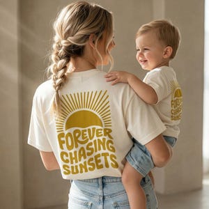 Könnte beinhalten: Cremefarbene T-Shirts mit goldenem Sonnendesign und dem Aufdruck "FOREVER CHASING SUNSETS" auf dem Rücken. Eine Person hält ein Kind, das ein passendes Shirt trägt. Die Shirts werden mit blauem Denim kombiniert.