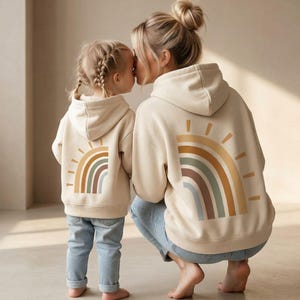 Könnte beinhalten: Zwei passende beige Kapuzenpullover mit einem Regenbogen-Sonnendesign auf dem Rücken. Eine Mutter und ihr Kind tragen die Kapuzenpullover, kombiniert mit hellblauen Jeans. Die Mutter kniet und küsst das Kind.