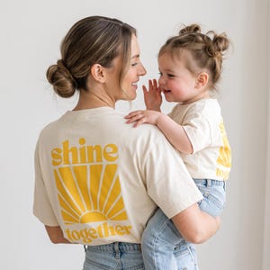 K&ouml;nnte beinhalten: Zwei cremefarbene T-Shirts mit kurzen &Auml;rmeln und einer gelben Grafik auf der R&uuml;ckseite. Die Grafik zeigt ein stilisiertes Sonnenmotiv mit dem Text "shine" &uuml;ber der Sonne und "together" darunter in einer fetten, abgerundeten Schrift.