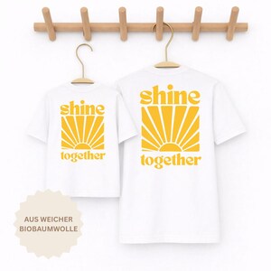 K&ouml;nnte beinhalten: Zwei wei&szlig;e T-Shirts an Holzkleiderb&uuml;geln, die jeweils mit einer gelben Grafik eines Sonnenstrahlendesigns und dem Text "shine together" in einer fetten, abgerundeten Schrift bedruckt sind. Die T-Shirts sind vor einem hellen Hintergrund.