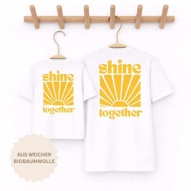 K&ouml;nnte beinhalten: Zwei wei&szlig;e T-Shirts an Holzkleiderb&uuml;geln, die jeweils mit einer gelben Grafik eines Sonnenstrahlendesigns und dem Text "shine together" in einer fetten, abgerundeten Schrift bedruckt sind. Die T-Shirts sind vor einem hellen Hintergrund.