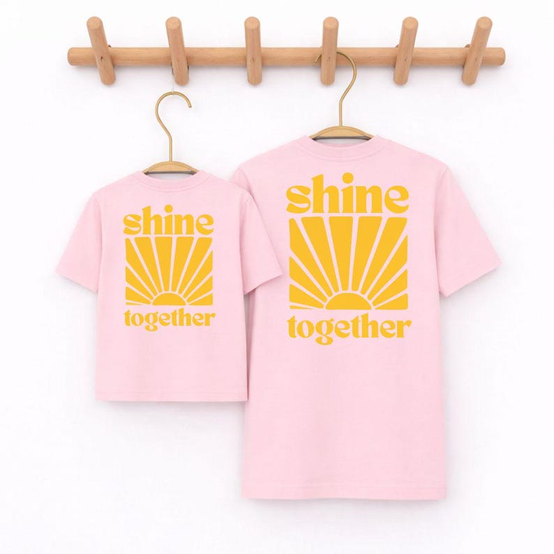 K&ouml;nnte beinhalten: Zwei hellrosa T-Shirts mit kurzen &Auml;rmeln, die an Holzkleiderb&uuml;geln h&auml;ngen. Jedes Shirt zeigt einen gelben Grafikdruck eines Sonnenaufgangs &uuml;ber dem Text "shine together" in einer fetten, abgerundeten, kleingeschriebenen gelben Schrift.