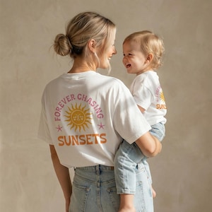 Könnte beinhalten: Weisse T-Shirts mit passendem "Forever Chasing Sunsets"-Design. Das Design zeigt eine Sonnenillustration in Orange und Rosa mit Text in einem Kreis. Die Shirts werden von einer Person und einem Kind getragen, beide in Jeans.