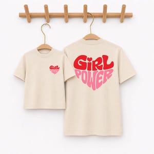 May include: Two beige short-sleeved t-shirts hanging on wooden hangers. The smaller shirt features a small red and pink heart graphic with the text Girl Power on the chest. The larger shirt displays a large heart graphic with Girl Power text.