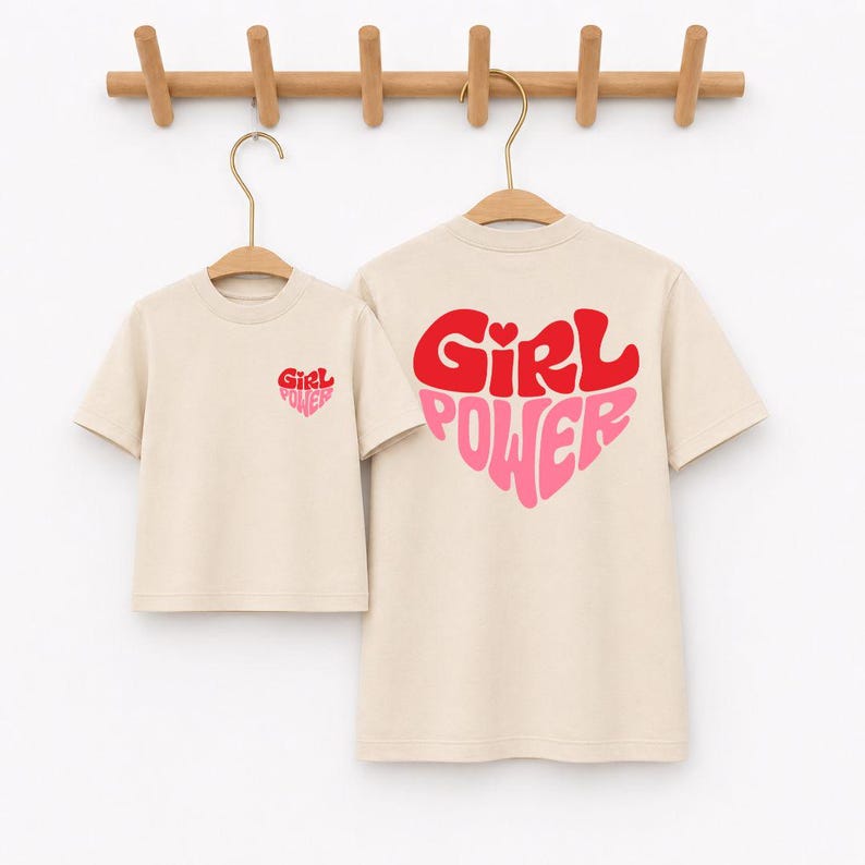 May include: Two beige short-sleeved t-shirts hanging on wooden hangers. The smaller shirt features a small red and pink heart graphic with the text Girl Power on the chest. The larger shirt displays a large heart graphic with Girl Power text.