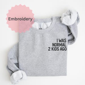 Könnte beinhalten: Ein meliertes graues Sweatshirt mit dem Text "I WAS NORMAL 2 KIDS AGO" in Schwarz. Das Wort "Embroidery" befindet sich in einer rosa Sternform oben links. Das Sweatshirt ist gefaltet, die Ärmel sind drapiert.