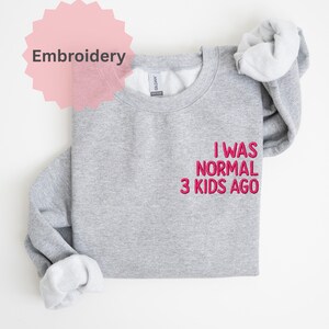 May include: A light grey sweatshirt with the embroidered text "I WAS NORMAL 3 KIDS AGO" in pink. The word "Embroidery" is in a pink starburst shape in the upper left corner. The sweatshirt has a crew neck and long sleeves.
