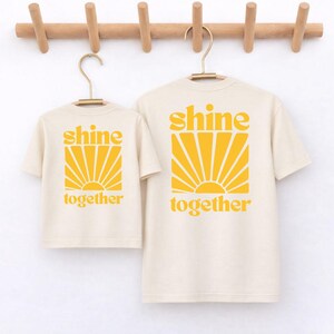 K&ouml;nnte beinhalten: Zwei cremefarbene T-Shirts mit kurzen &Auml;rmeln in verschiedenen Gr&ouml;&szlig;en, jeweils mit einem gelben Grafikdruck eines Sonnenstrahlen-Motivs und dem Text "shine together" in einer fetten, abgerundeten Schrift auf der Vorderseite.