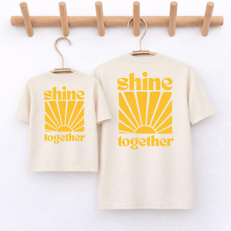 K&ouml;nnte beinhalten: Zwei cremefarbene T-Shirts mit kurzen &Auml;rmeln in verschiedenen Gr&ouml;&szlig;en, jeweils mit einem gelben Grafikdruck eines Sonnenstrahlen-Motivs und dem Text "shine together" in einer fetten, abgerundeten Schrift auf der Vorderseite.