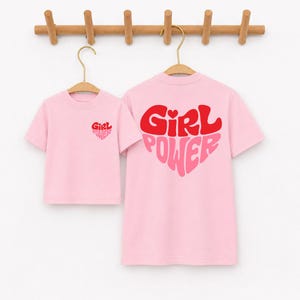 May include: Two light pink short-sleeve t-shirts hanging on wooden hangers. The smaller shirt features a small red and pink heart graphic with the text Girl Power on the chest. The larger shirt displays a large heart graphic with Girl Power text.
