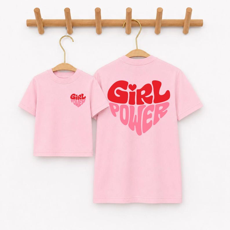 May include: Two light pink short-sleeve t-shirts hanging on wooden hangers. The smaller shirt features a small red and pink heart graphic with the text Girl Power on the chest. The larger shirt displays a large heart graphic with Girl Power text.