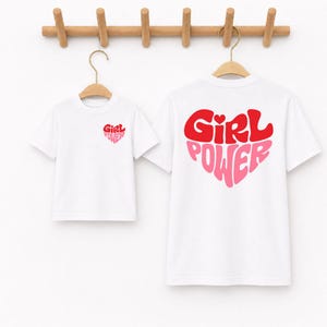 May include: Two white short-sleeved t-shirts hanging on wooden hangers. The smaller shirt features a small red and pink heart graphic with the text "Girl Power" on the chest. The larger shirt displays a large heart graphic with "Girl Power" text.