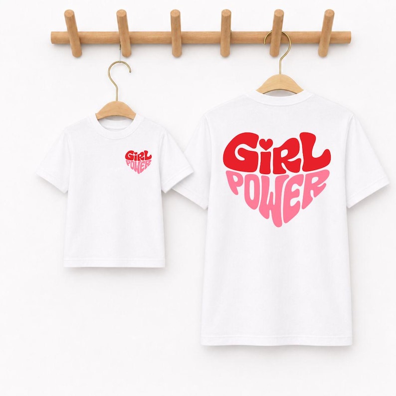 May include: Two white short-sleeved t-shirts hanging on wooden hangers. The smaller shirt features a small red and pink heart graphic with the text "Girl Power" on the chest. The larger shirt displays a large heart graphic with "Girl Power" text.