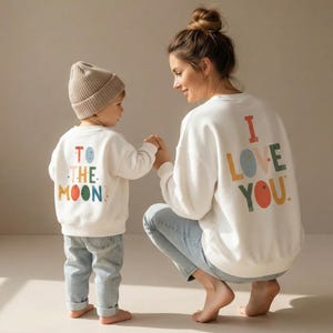 Puede incluir: Dos sudaderas blancas con texto colorido. Una sudadera dice "I LOVE YOU" y la otra "TO THE MOON!" Un niño lleva un gorro beige y vaqueros azul claro. El adulto lleva vaqueros azul claro.